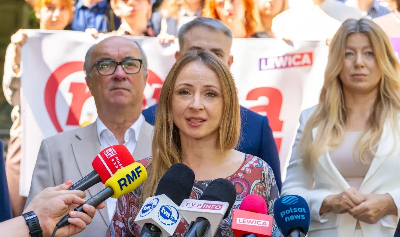 Kobieta przemawia do mediów podczas konferencji prasowej, otoczona licznymi mikrofonami, a w tle widać inne osoby i baner Lewicy.
