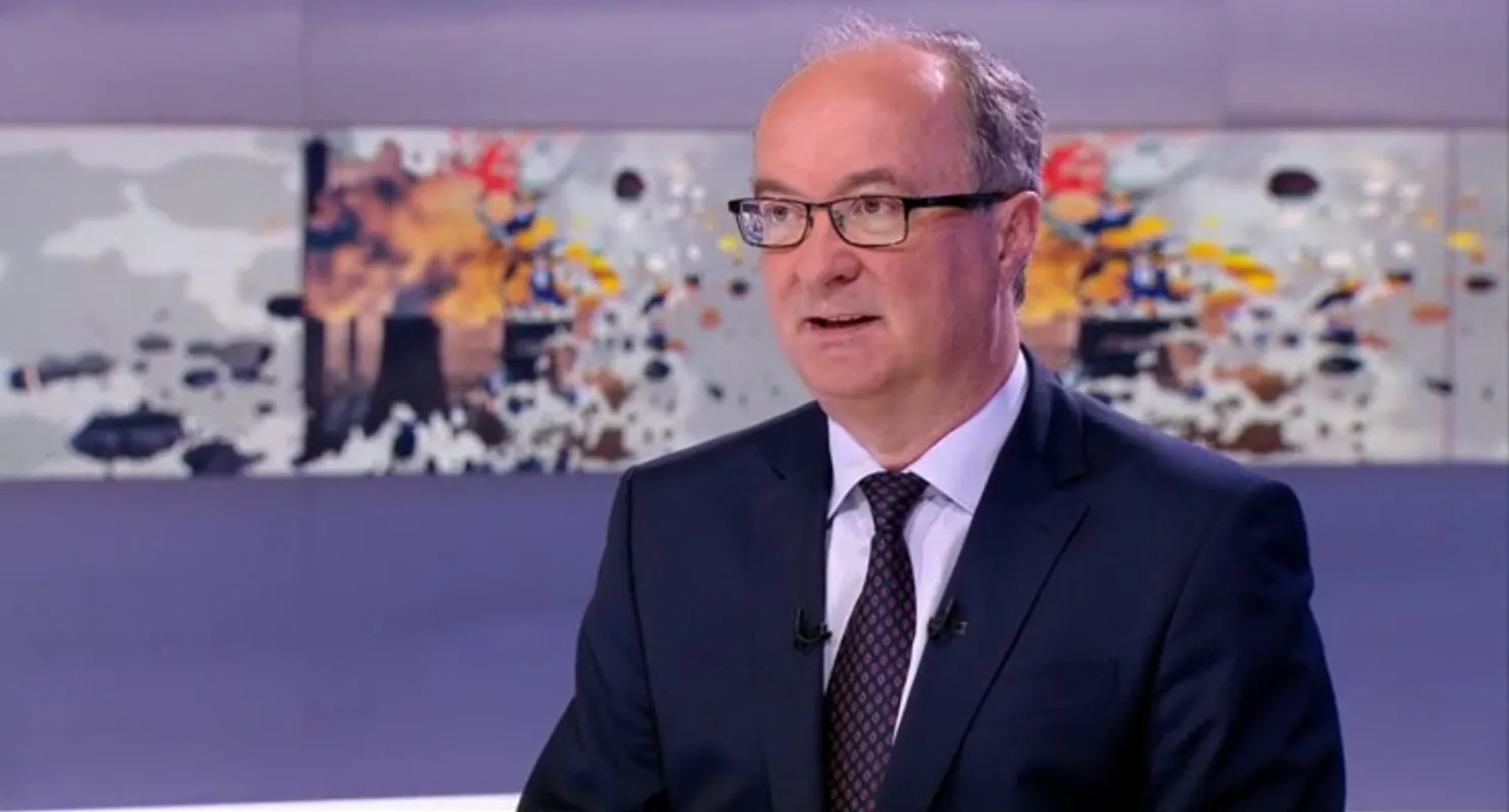 Mężczyzna w garniturze i okularach siedzi w studiu telewizyjnym i rozmawia, a w tle widać mapę świata.