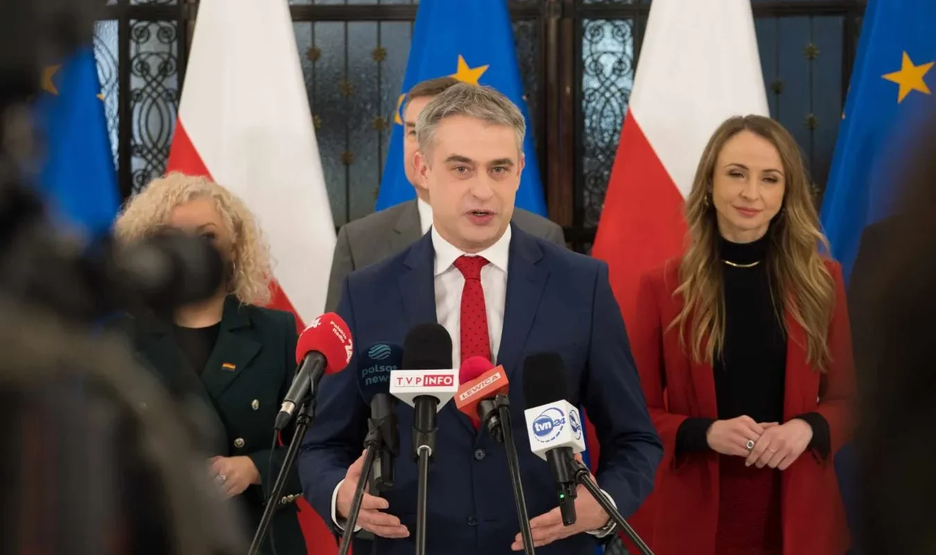 Polityk w granatowym garniturze wygłasza oświadczenie podczas konferencji prasowej z licznymi mikrofonami, stojąc na tle flag Polski i Unii Europejskiej, w kontekście sporu o Starlinki.