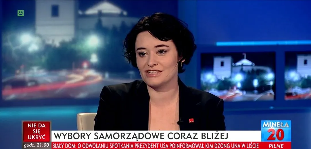 Kobieta z krótkimi czarnymi włosami w czarnej marynarce siedzi w studiu telewizyjnym, ma na klapie czerwony pin, a na dole ekranu przewijają się napisy informacyjne.