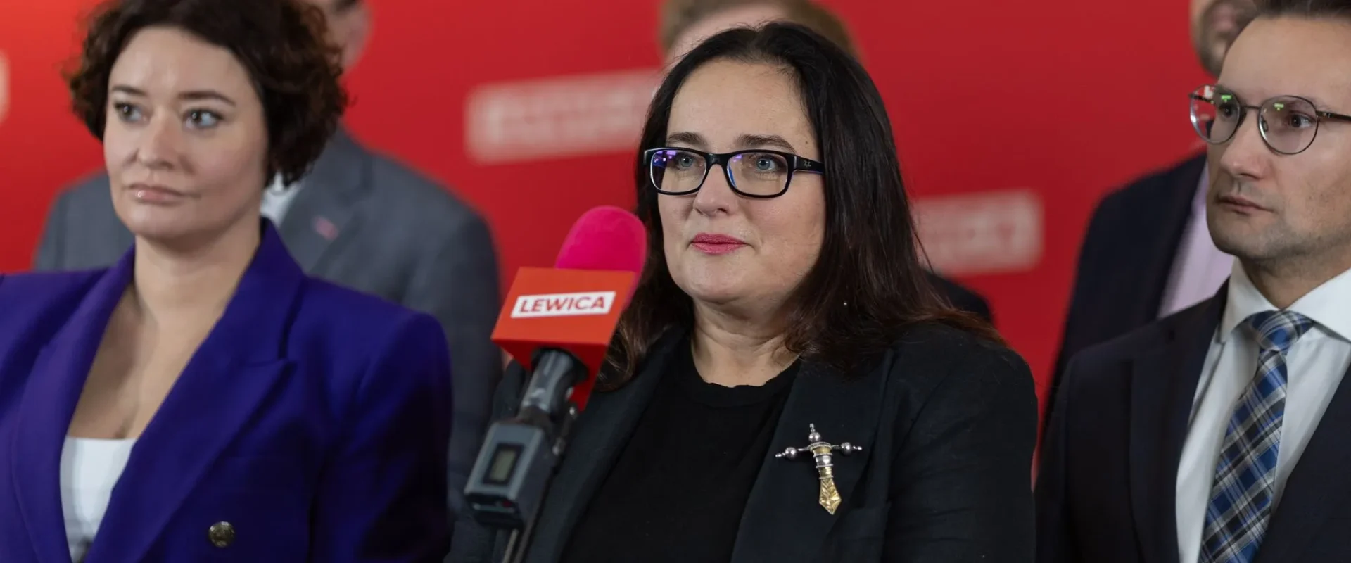 Kobieta w czarnym ubraniu i czarnych okularach przemawia do mikrofonu z napisem Lewica podczas konferencji prasowej, otoczona przez dwie osoby na czerwonym tle.