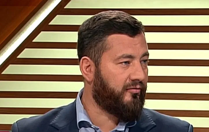 Mężczyzna z brodą w granatowym garniturze siedzi w studiu telewizyjnym na tle drewnianych paneli.