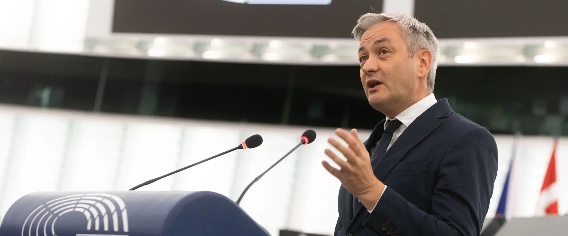 Polski polityk w garniturze przemawia z mównicy w sali plenarnej Parlamentu Europejskiego, na tle symbolu Unii Europejskiej.