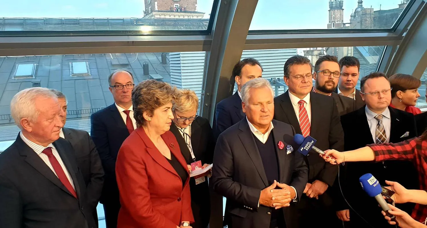 Grupa polityków w garniturach i dziennikarzy na konferencji prasowej w jasnym, nowoczesnym wnętrzu z dużymi oknami i mikrofonami skierowanymi do mówcy.