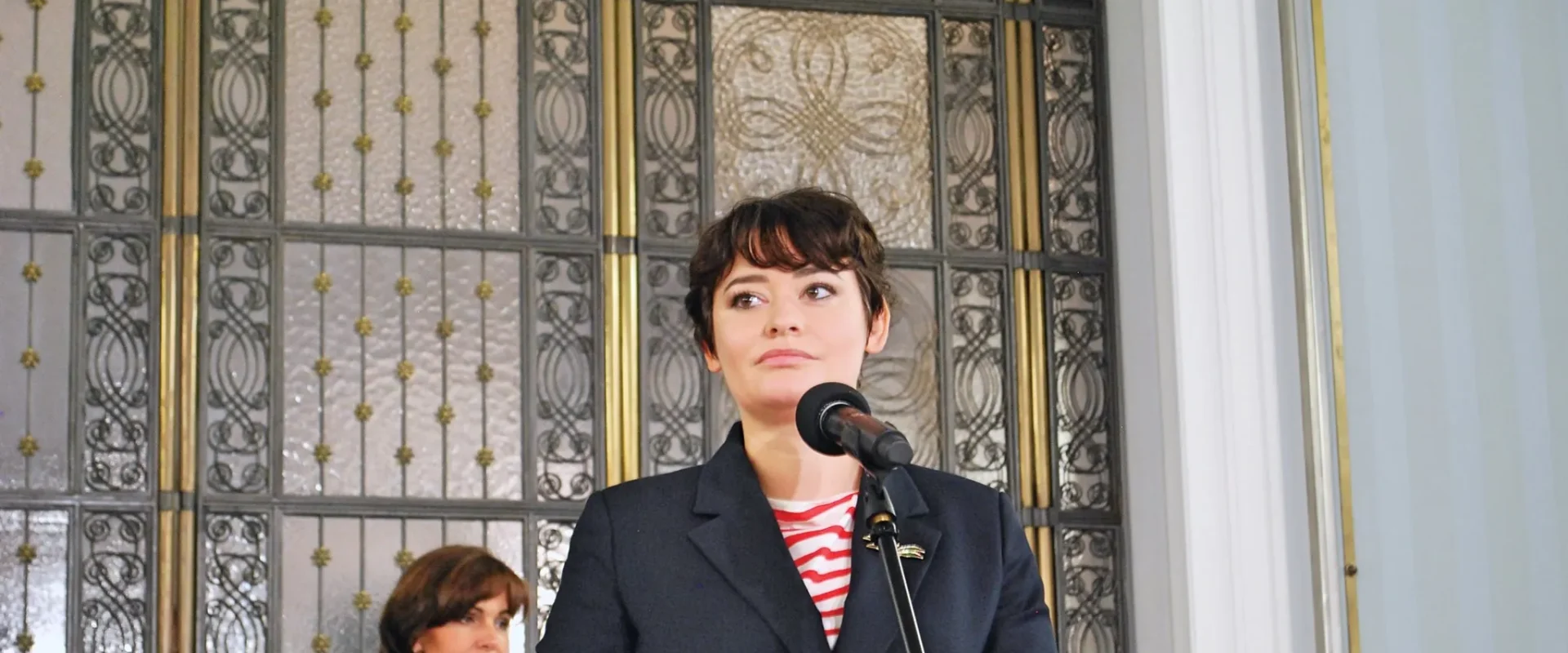 Kobieta w ciemnym garniturze przemawia do mikrofonu podczas konferencji; w tle widoczne ozdobne, metalowe drzwi, a za nią stoi druga kobieta.