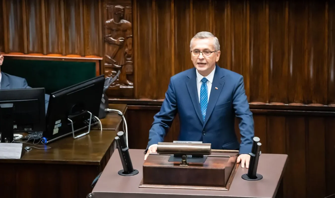 Mężczyzna w garniturze przemawia w sali obrad, w tle widoczne drewniane elementy dekoracyjne.