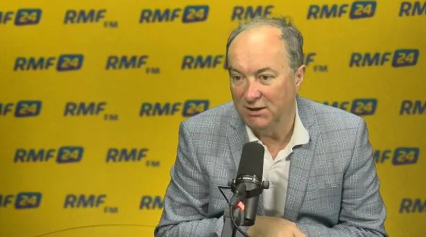 Mężczyzna w kratkowanej marynarce i białej koszuli siedzi przy mikrofonie w studiu RMF FM, na żółtym tle z powtarzającym się logiem RMF24.