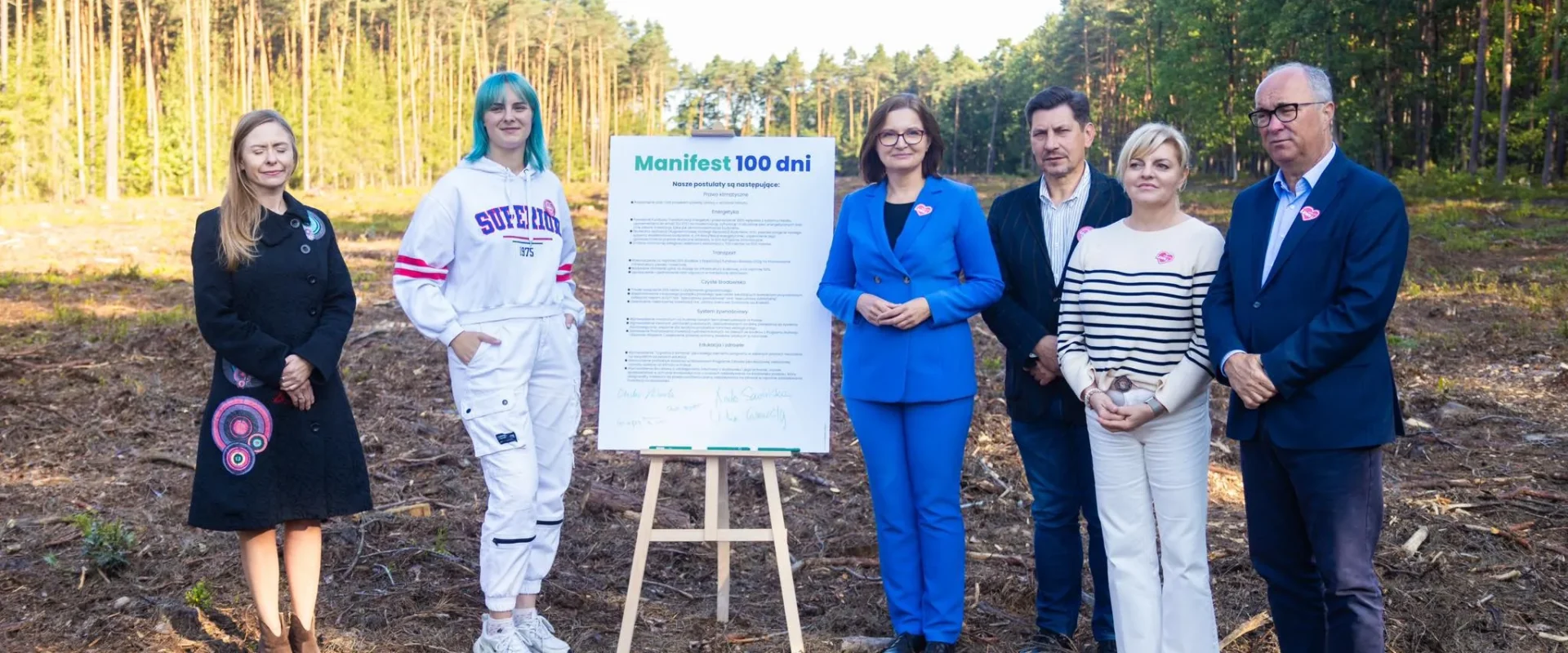 Grupa sześciu osób stoi na leśnej polanie z lasem w tle, obok tablicy z napisem „Manifest 100 dni dla polskiej przyrody i klimatu”.