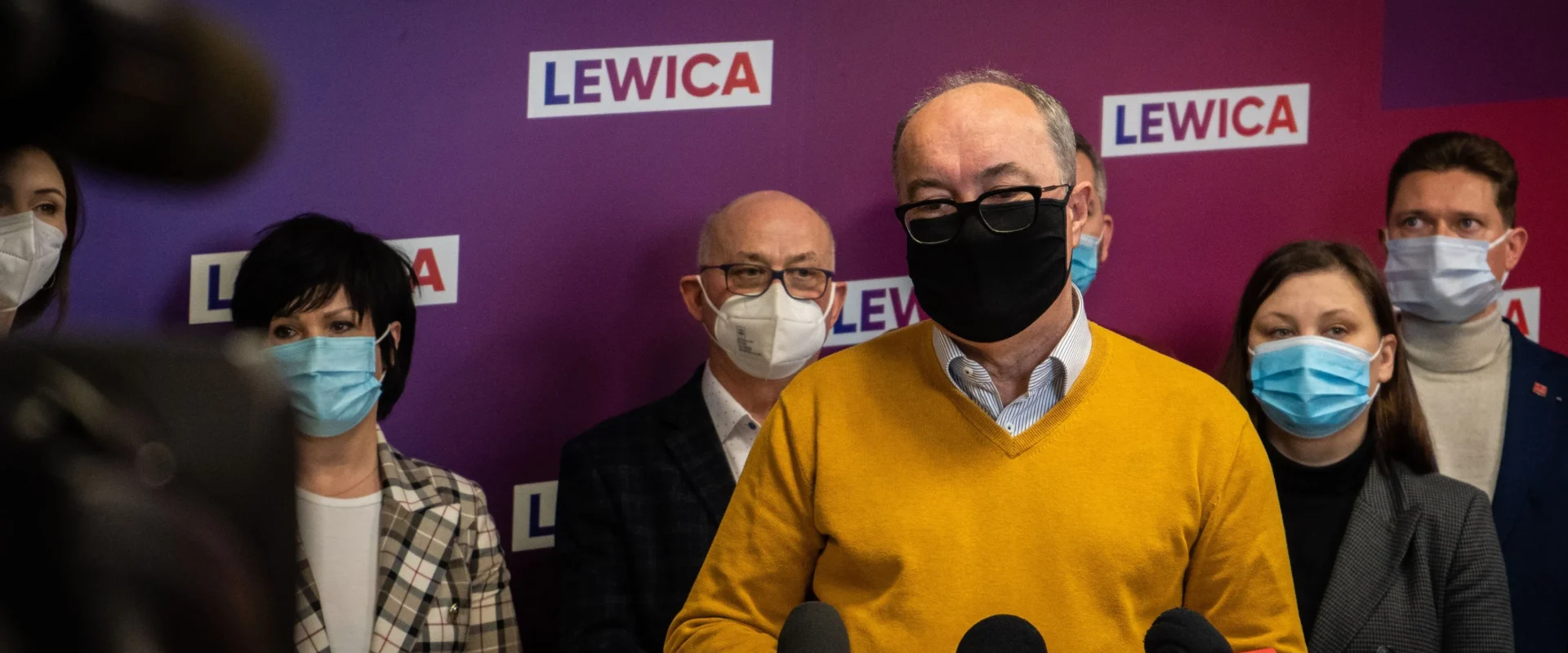 Na zdjęciu widać mężczyznę w żółtej bluzie, otoczonego przez grupę osób w maseczkach, podczas konferencji prasowej przed tłem z logo partii Lewica.