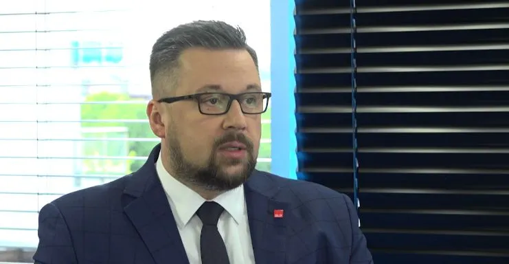 Mężczyzna w garniturze i okularach siedzi w studiu, mówiąc do kamery, a w tle widoczne są żaluzje i czarne rolety.