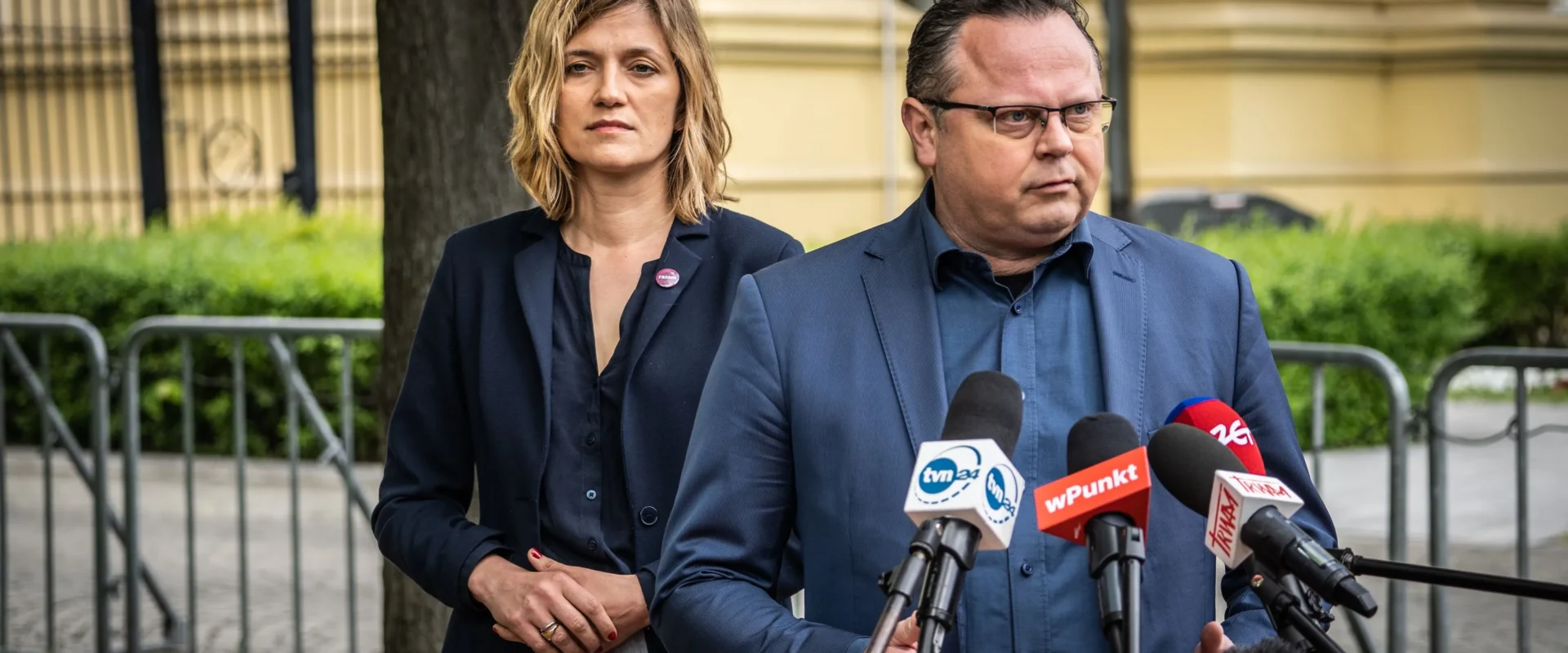 Mężczyzna w niebieskiej marynarce przemawia do mediów podczas konferencji prasowej, stojąc obok kobiety w granatowej marynarce, przed mikrofonami.