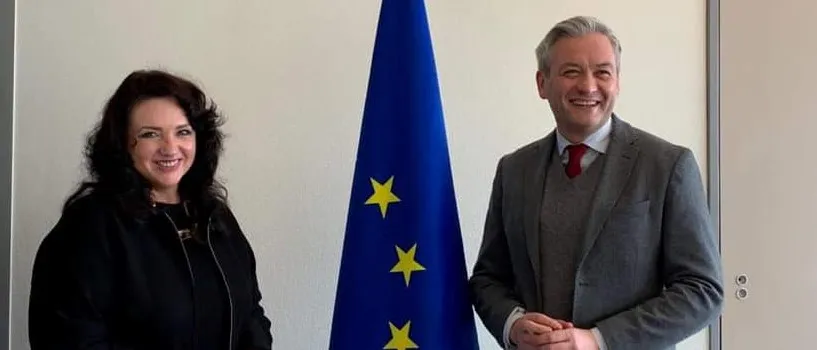 Dwoje dorosłych w formalnych strojach stoi obok siebie przy fladze Unii Europejskiej, oboje uśmiechnięci.