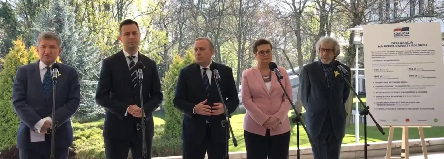 Grupa pięciu osób w garniturach stoi przy mikrofonach podczas konferencji prasowej na zewnątrz, obok plansza z napisem Deklaracja na rzecz oświaty polskiej.