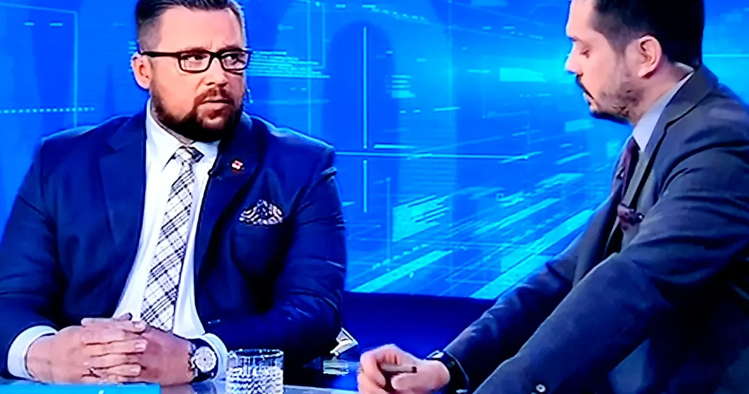 Dwóch mężczyzn rozmawia w studiu telewizyjnym na tle niebieskiej grafiki; po lewej w granatowym garniturze z kraciastym krawatem i okularami, po prawej w szarym garniturze, przy stole z notatkami i logiem programu „TWÓJ WYBÓR”.