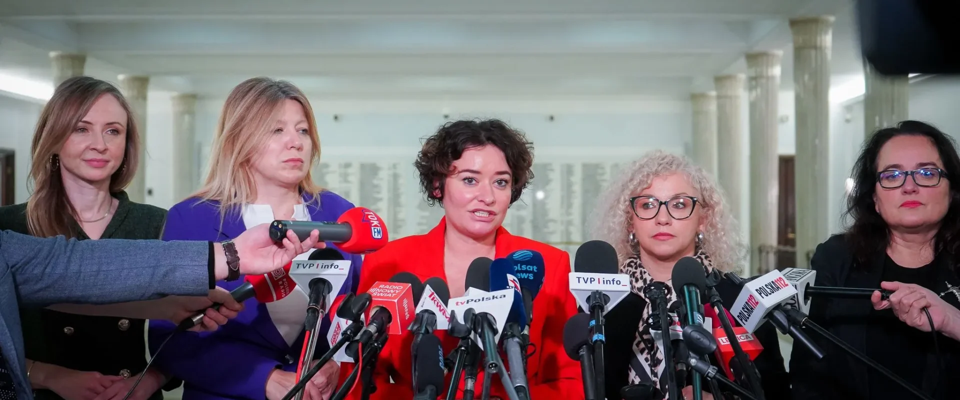 Grupa kobiet podczas konferencji prasowej, otoczona licznymi mikrofonami i kamerami, w biurowej przestrzeni z kolumnami w tle.