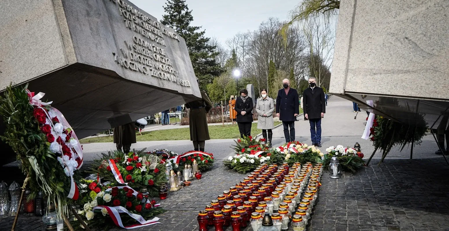 Grupa osób w maseczkach stoi w parku przed pomnikiem, upamiętniając 11. rocznicę katastrofy smoleńskiej, otoczona kwiatami i zapalonymi zniczami.