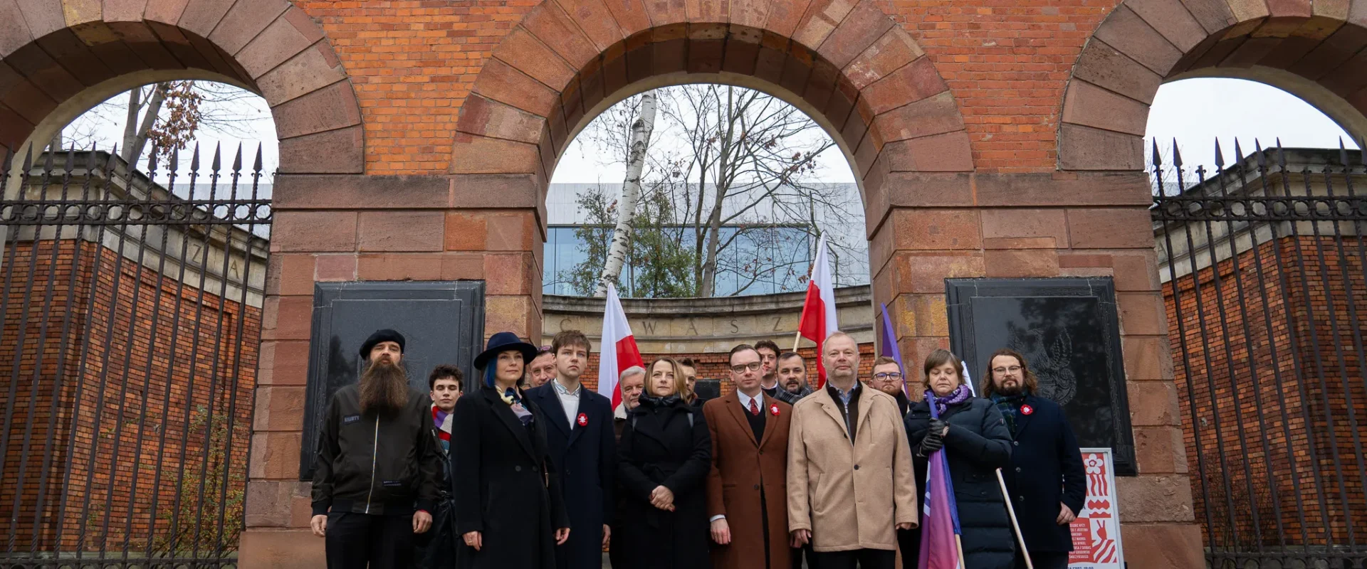 Grupa osób stoi przed ceglaną bramą z trzema łukami, trzymając polskie flagi podczas obchodów Narodowego Święta Niepodległości.