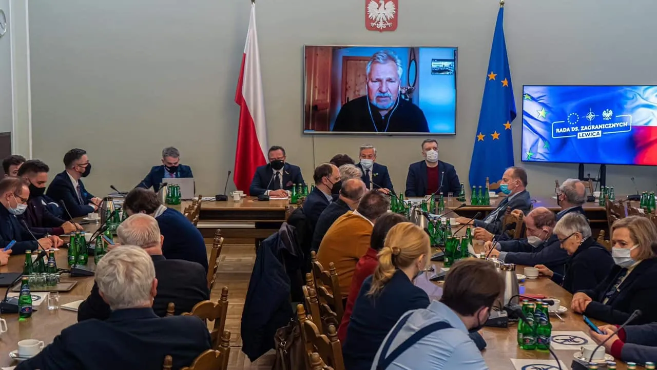 Na zdjęciu przedstawiona jest sala konferencyjna, w której odbywa się spotkanie, a w tle widoczny jest ekran z osobą uczestniczącą w rozmowie online.