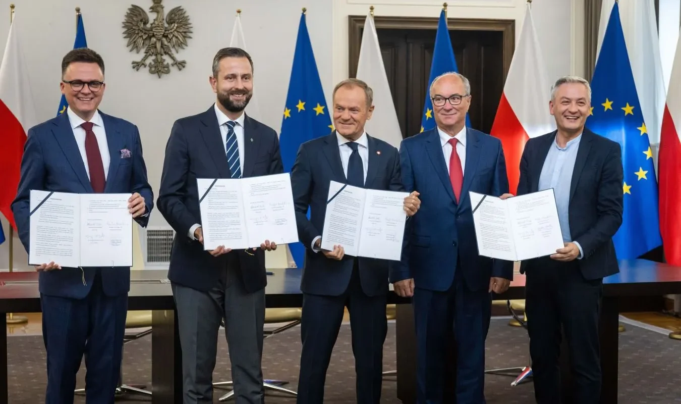 Pięciu mężczyzn w garniturach trzyma podpisane dokumenty, stojąc przed flagami Polski i Unii Europejskiej podczas wydarzenia koalicji szerokiego kompromisu.