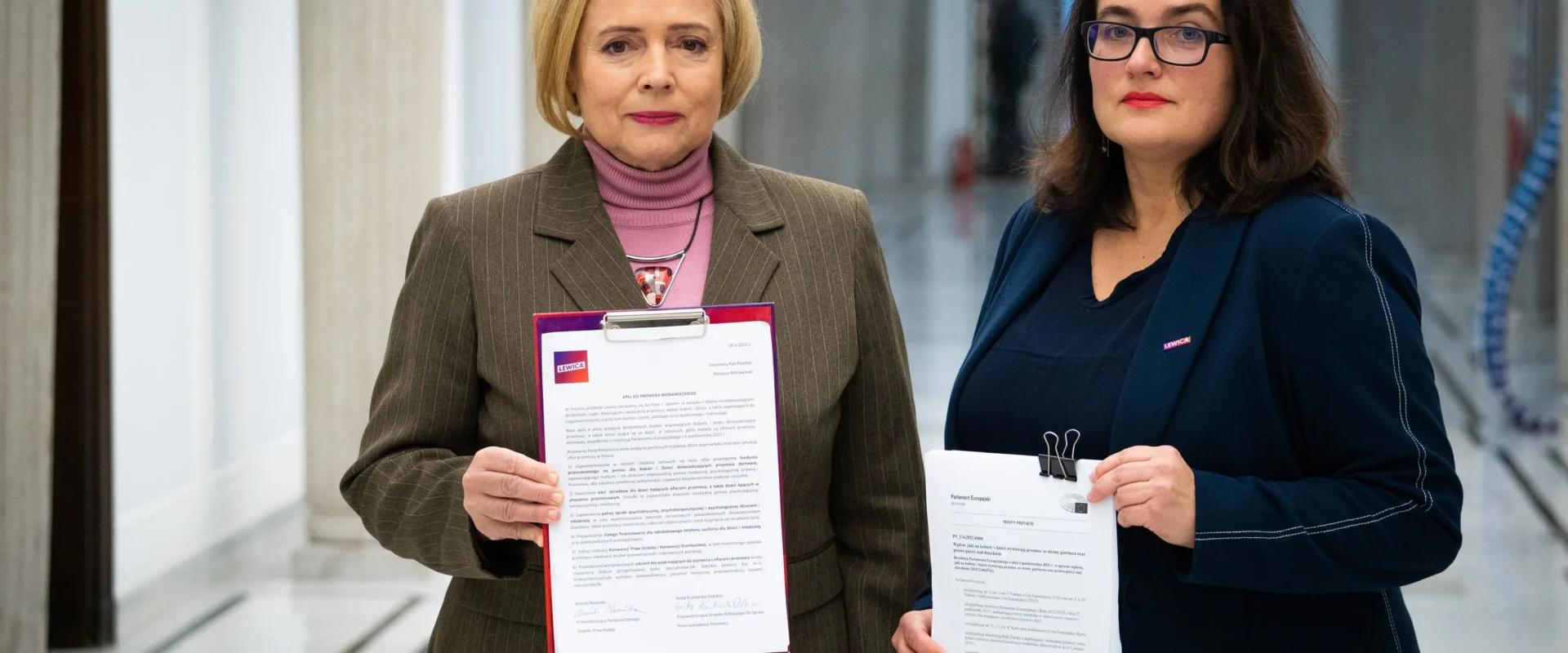 Na zdjęciu dwie kobiety stoją w korytarzu, trzymając dokumenty, z wyraźnym logo Lewicy na jednej z nich.