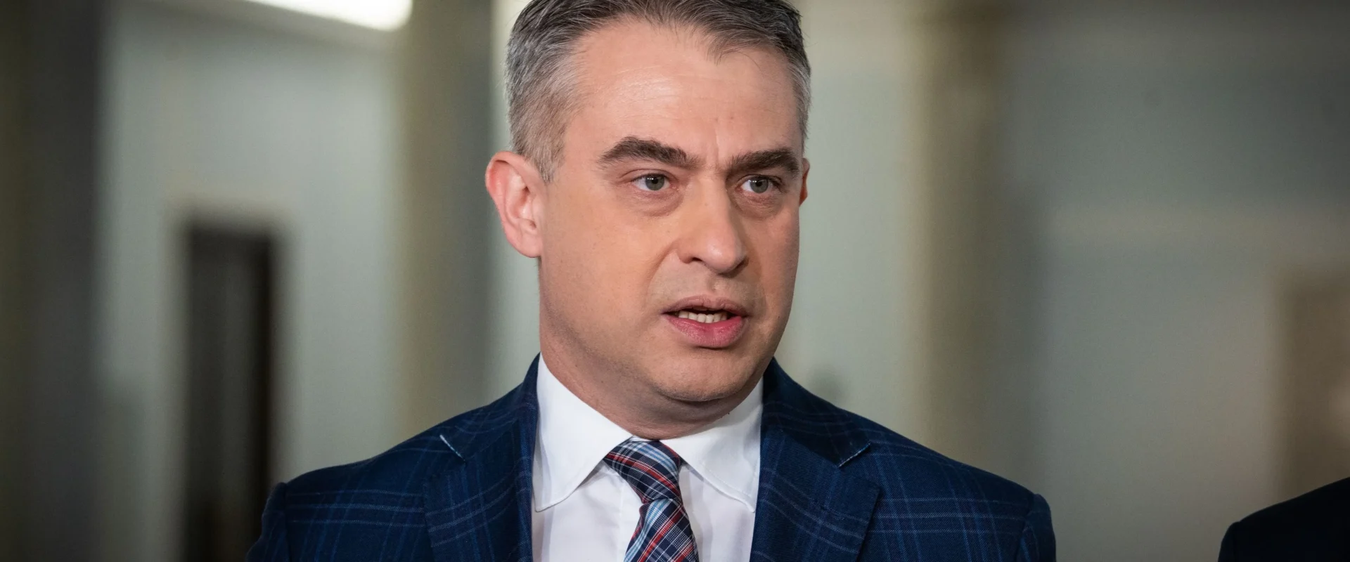 Mężczyzna w granatowym garniturze z kraciastym krawatem przemawia do dziennikarzy podczas konferencji prasowej, stojąc przed ustawionymi mikrofonami.