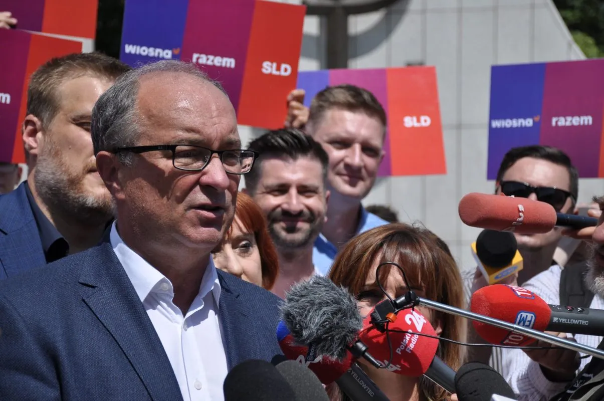Politycy koalicji Lewicy SLD, Wiosna i Razem wygłaszają oświadczenie podczas konferencji prasowej, otoczeni przez dziennikarzy i mikrofony z plakatami w kolorach partii w tle.