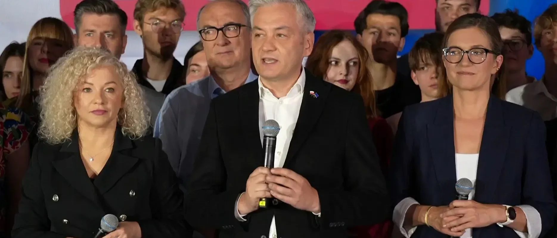 Trzy osoby stojące na scenie trzymające mikrofony podczas przemówienia, w tle zgromadzona publiczność.
