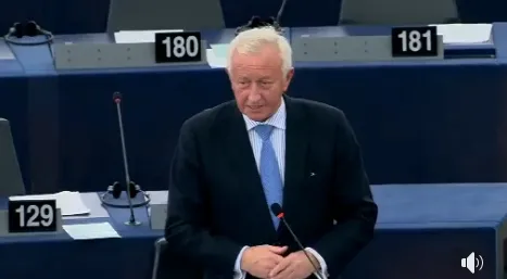 Mężczyzna w ciemnym garniturze przemawia do mikrofonu podczas sesji parlamentu.
