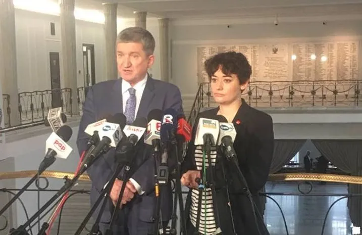 Dwójka polityków stoi przed rzędem mikrofonów podczas konferencji prasowej w holu parlamentu.