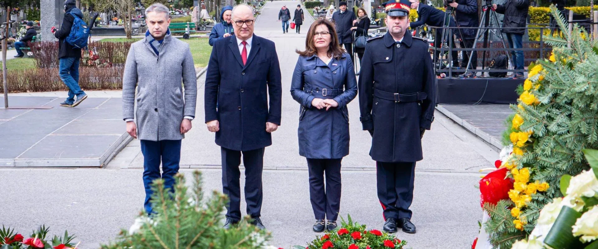 Czterech ludzi stoi w parku otoczonych kwiatami i wieńcami upamiętniającymi 12. rocznicę tragedii smoleńskiej, przy obecności kamer i fotoreporterów w tle.