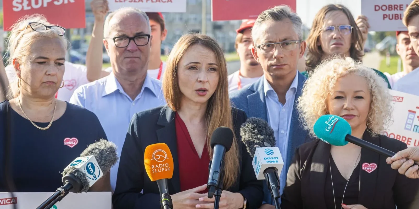 Grupa polityków na konferencji prasowej, pośrodku kobieta przemawia do mikrofonów, w tle banery Lewicy i zgromadzeni dziennikarze.