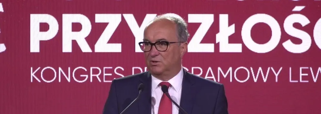 Mężczyzna w garniturze i czerwonym krawacie przemawia z podium na tle czerwonej sceny z dużymi białymi napisami.