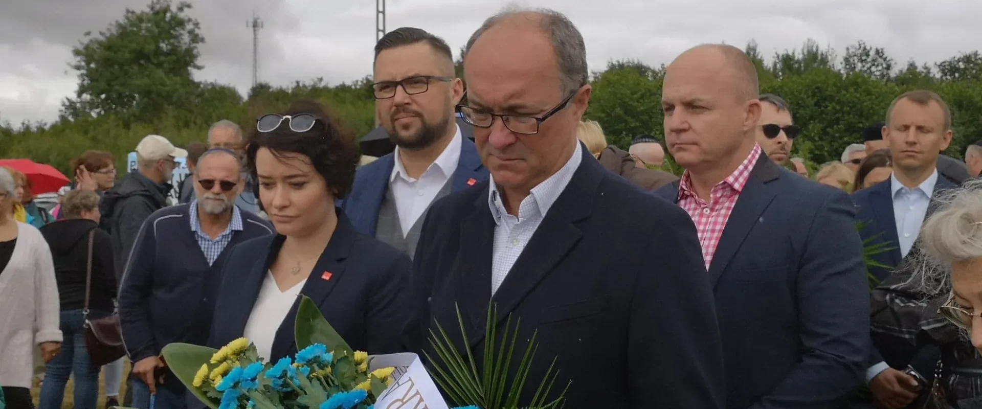 Grupa osób w formalnych ubraniach składa wieńce i kwiaty podczas uroczystości upamiętniającej zamordowanych Żydów w Jedwabnem.