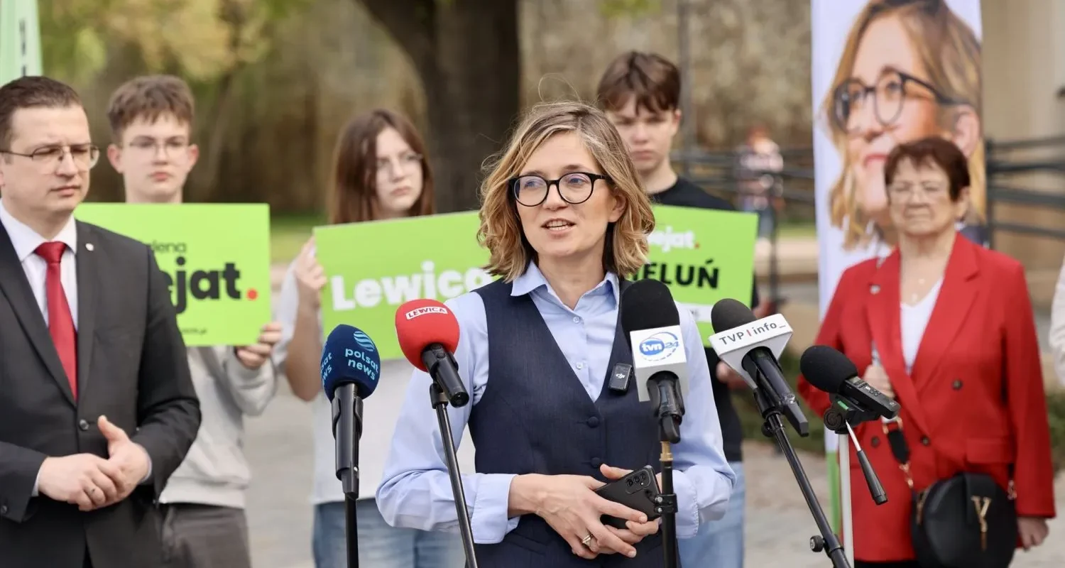 Polityczka z okularami przemawia na konferencji prasowej przed mikrofonami, w tle stoją ludzie trzymający zielone plakaty, a kontekst dotyczy ograniczenia wpływu korporacji na prawo.