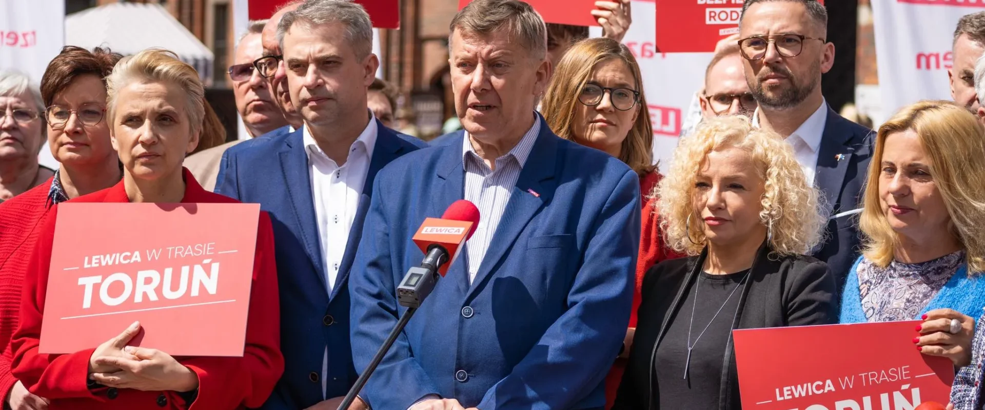 Grupa osób w formalnych ubraniach stoi na konferencji prasowej, trzyma plakaty z napisem "Lewica w trasie Toruń" i przemawia do mikrofonu.
