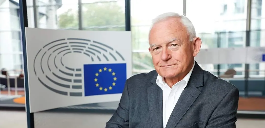 Starszy mężczyzna w białej koszuli i szarym garniturze stoi w biurze, a w tle widoczne jest logo Unii Europejskiej.