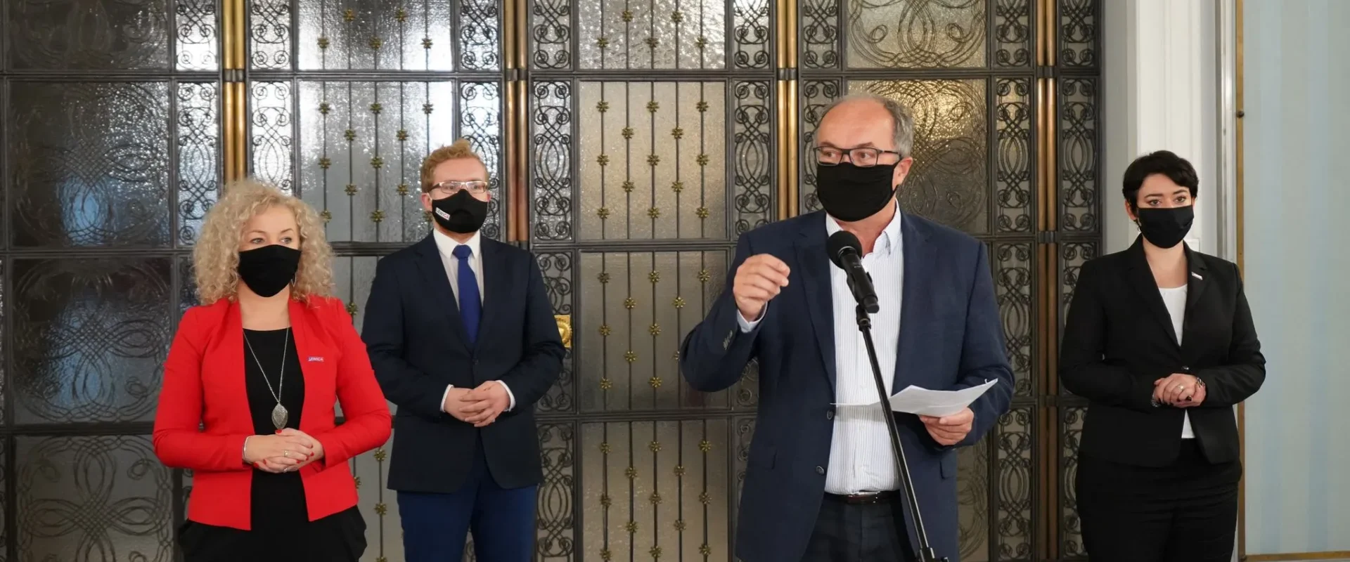 Czterech polityków w maseczkach stoi przy mikrofonie podczas konferencji prasowej, na tle ozdobnych metalowych drzwi.
