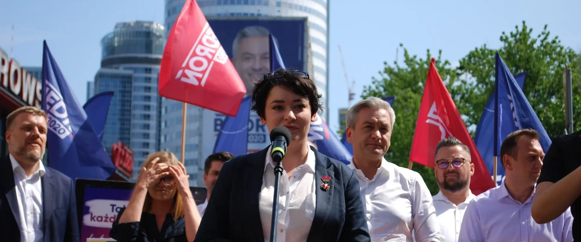 Kobieta w garniturze przemawia do zgromadzonej publiczności z mikrofonem podczas plenerowego zgromadzenia, wokół niej stoją ludzie z flagami, a w tle widać wieżowiec.