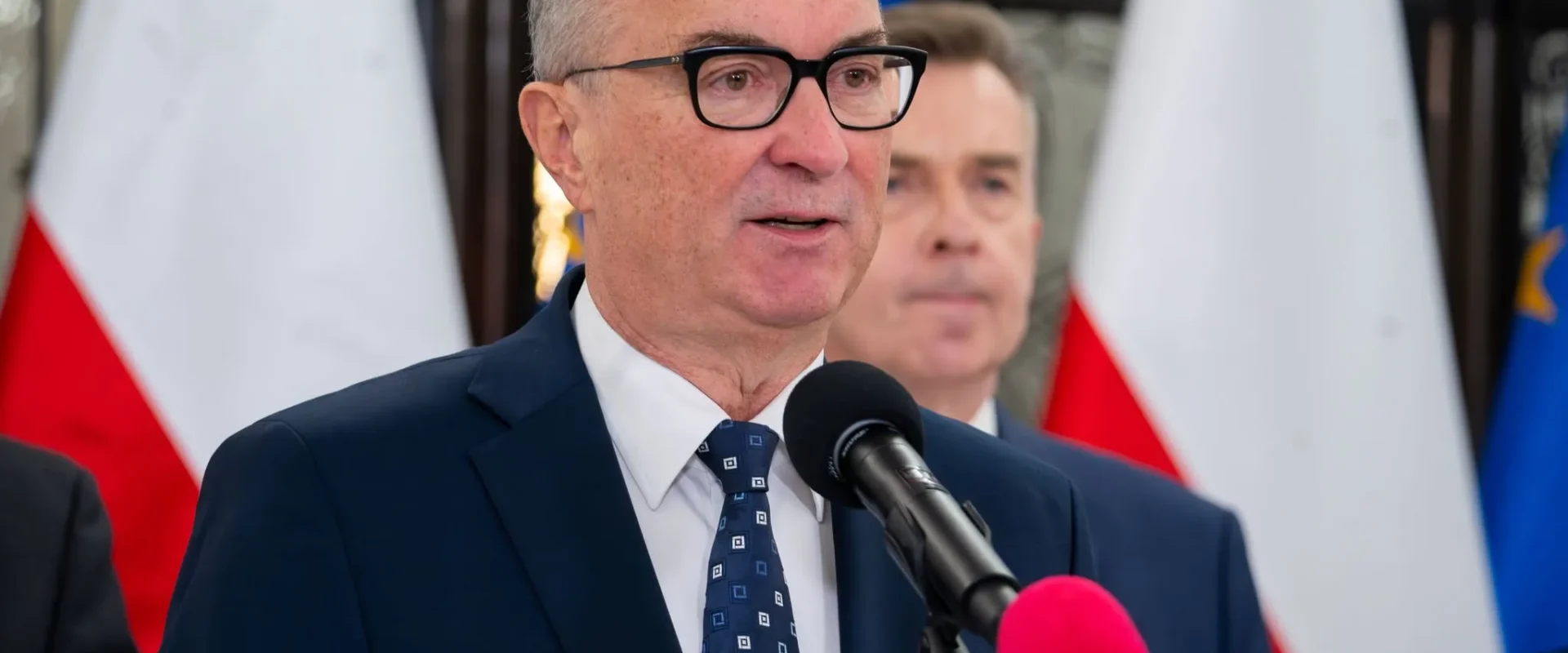 Mężczyzna w garniturze i okularach przemawia do mikrofonu podczas konferencji prasowej, z flagami w tle.