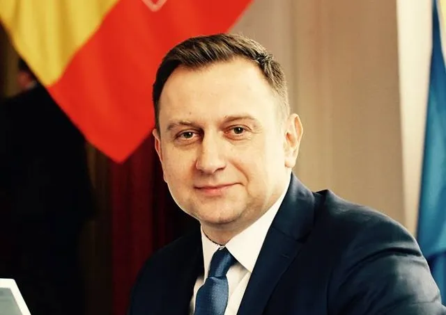 Mężczyzna w garniturze siedzi przy stole, na tle widoczne flagi, uśmiecha się do kamery.