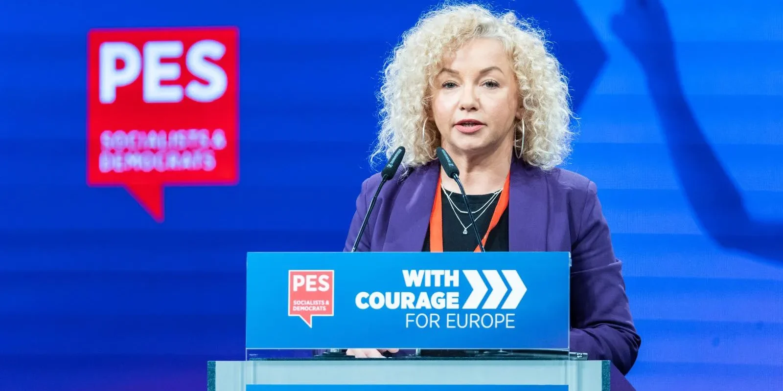 Kobieta o kręconych blond włosach w fioletowym żakiecie stoi za mównicą z logiem PES i napisem „With Courage for Europe”, przemawiając na niebieskiej scenie.