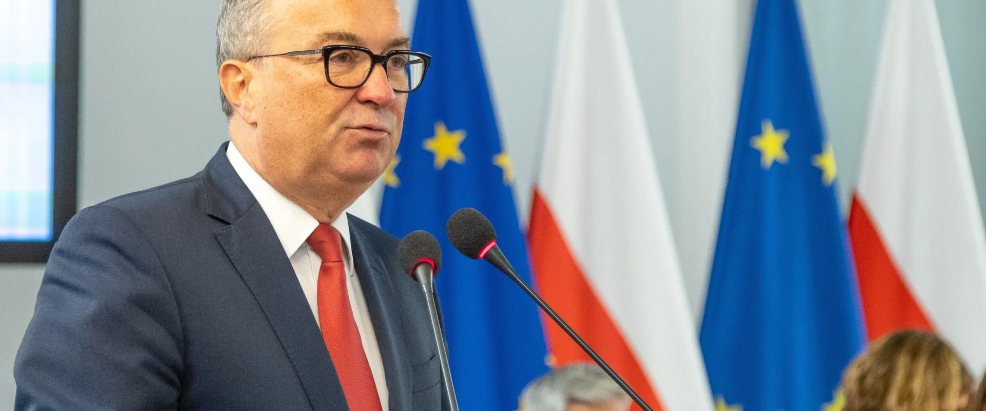 Mężczyzna w garniturze mówi do mikrofonu na tle flag europejskich i polskich podczas konferencji.