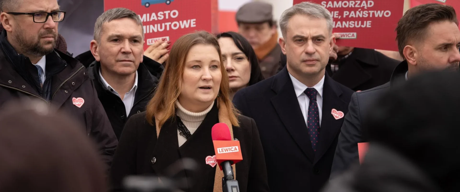 Kobieta przemawia do mikrofonu podczas wiecu Lewicy, otoczona przez mężczyzn i czerwone plakaty z logo Lewicy w tle.
