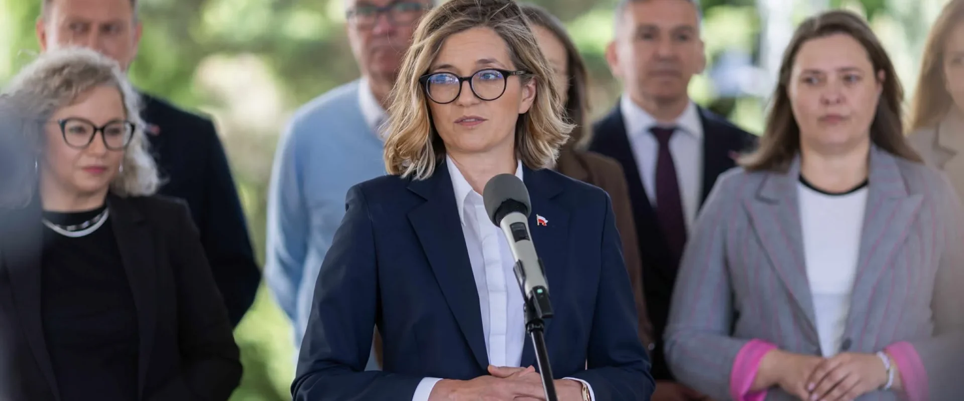 Kobieta w granatowym garniturze i okularach przemawia do mikrofonu podczas konferencji prasowej na zewnątrz, w tle stoi grupa współpracowników.