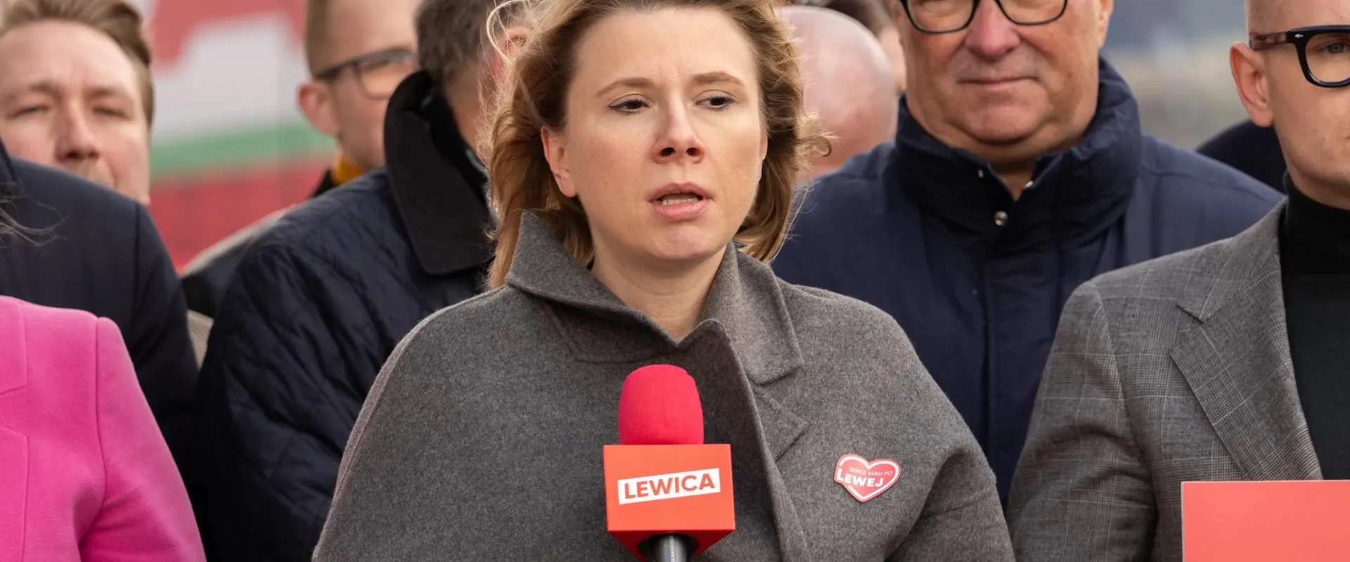 Kobieta przemawia do mikrofonu podczas konferencji, w otoczeniu innych osób; na czerwonym mikrofonie widnieje napis Lewica.