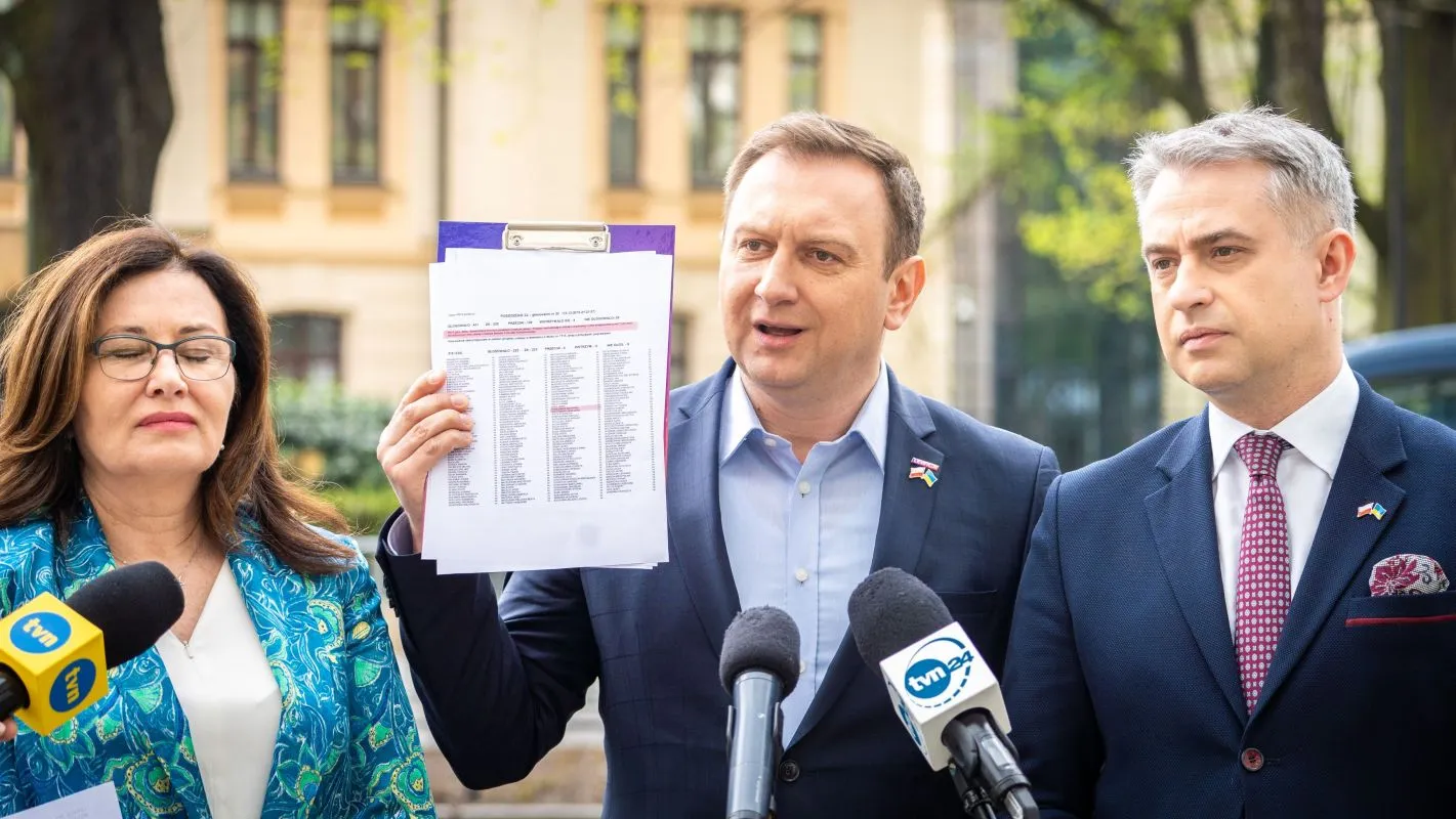Na zdjęciu trzech polityków uczestniczy w konferencji prasowej, podczas której jeden z nich trzyma dokument z wyraźnie widoczną listą, a obok znajdują się mikrofony mediów.