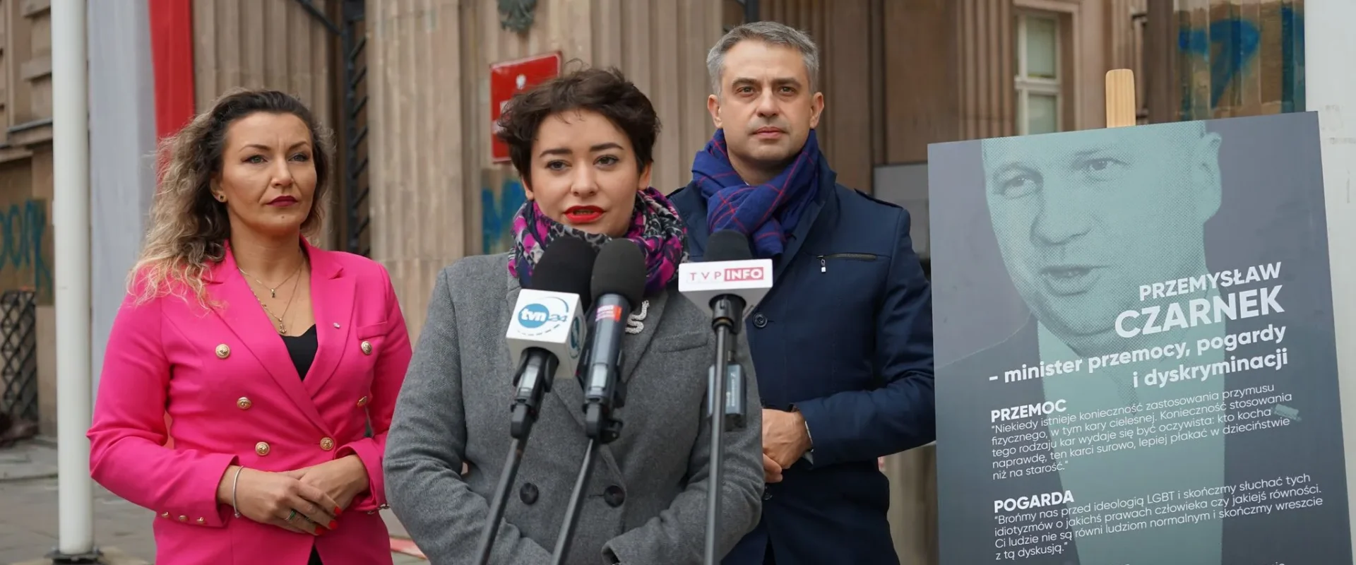 Trzy osoby stoją przed budynkiem podczas konferencji prasowej; kobieta w szarym płaszczu mówi do mikrofonów ustawionych na stojakach, a po prawej widoczny jest plakat z wizerunkiem mężczyzny i tekstem.