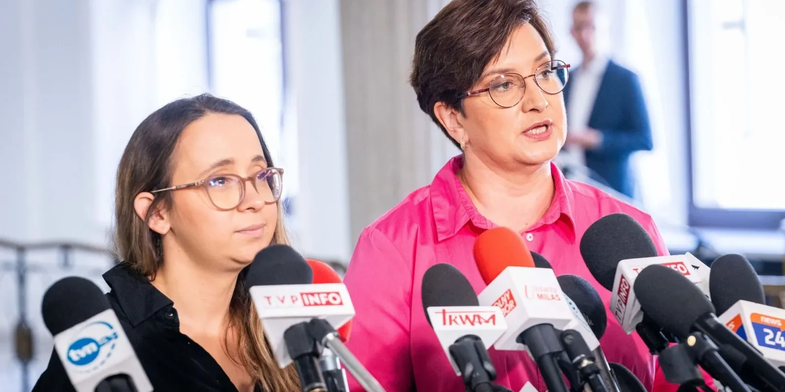 Dwie kobiety podczas konferencji prasowej stoją za licznymi mikrofonami i udzielają wypowiedzi mediom.
