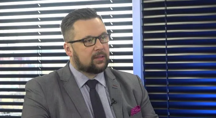Mężczyzna w szarym garniturze, ciemnym krawacie i różowej poszetce, w okularach i z brodą, siedzi na tle poziomych żaluzji.