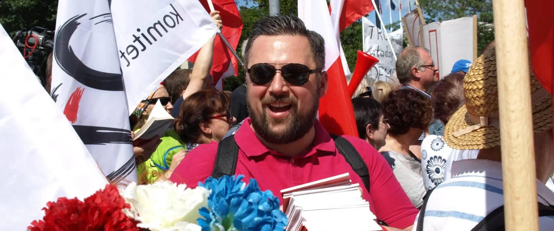 Uśmiechnięty mężczyzna w różowym polo i okularach przeciwsłonecznych stoi w tłumie na demonstracji z flagami Polski, trzymając stos czerwono-białych broszur.